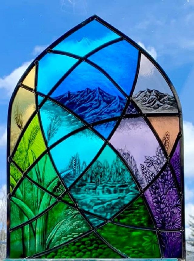 Colourful stained glass panel with mountains, plants and river scenes against blue sky.