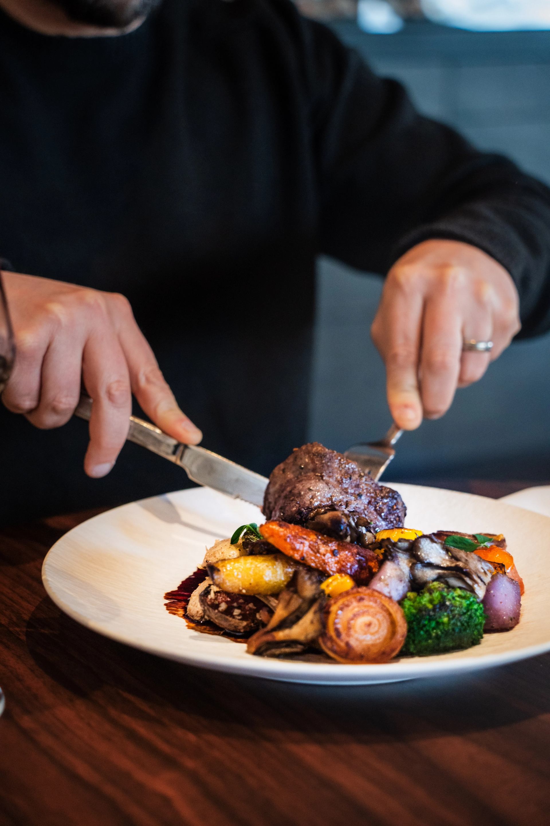 Person cuts cooked meat with roasted vegetables on a white plate at wooden table.