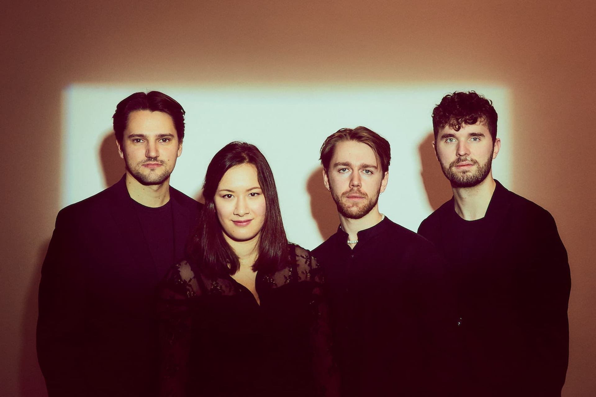 Four people in dark clothing stand against a wall with a bright rectangular light.