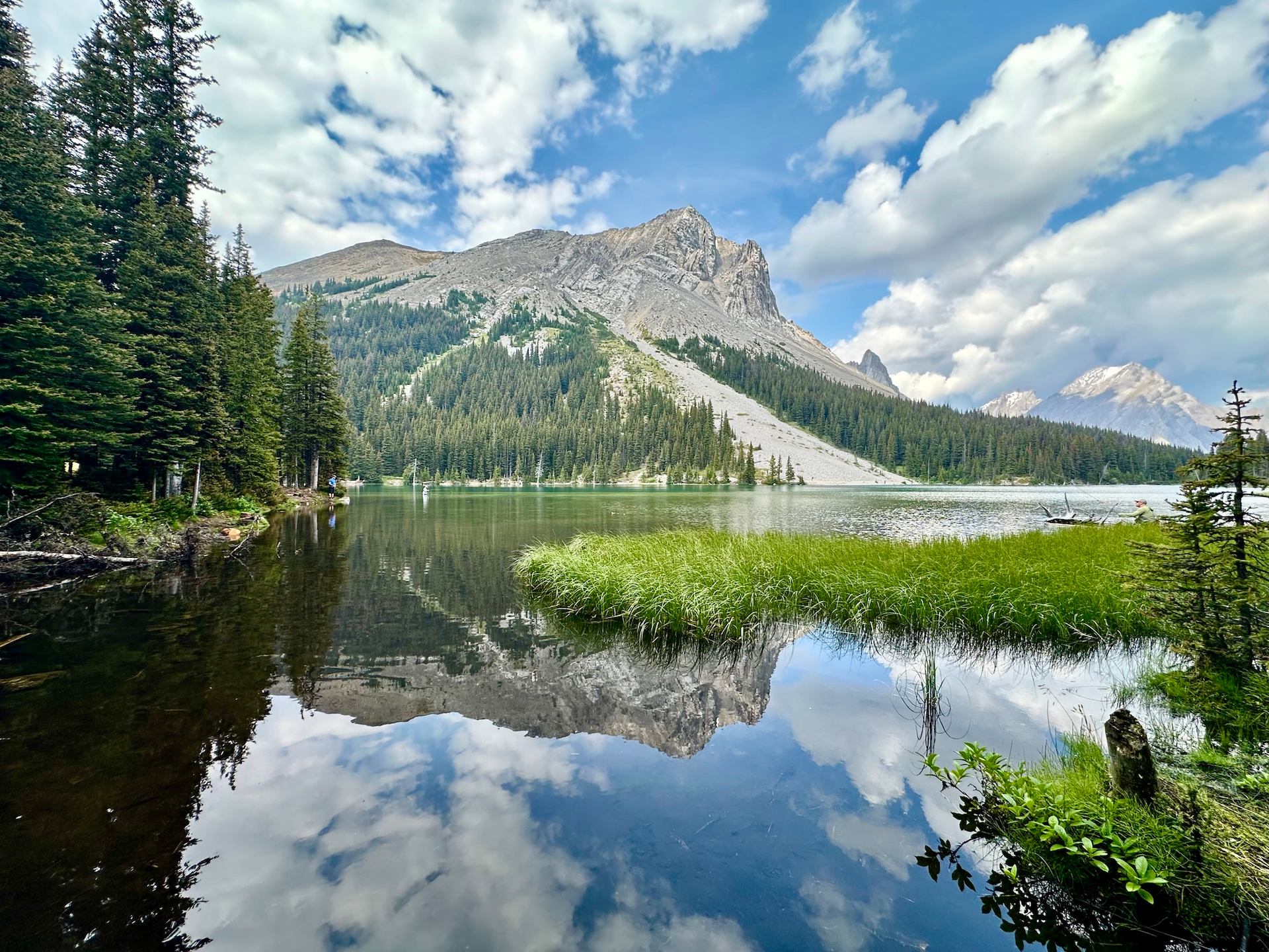 Mountain lake with evergreen forest, grassy shoreline and clouds reflected in calm water.