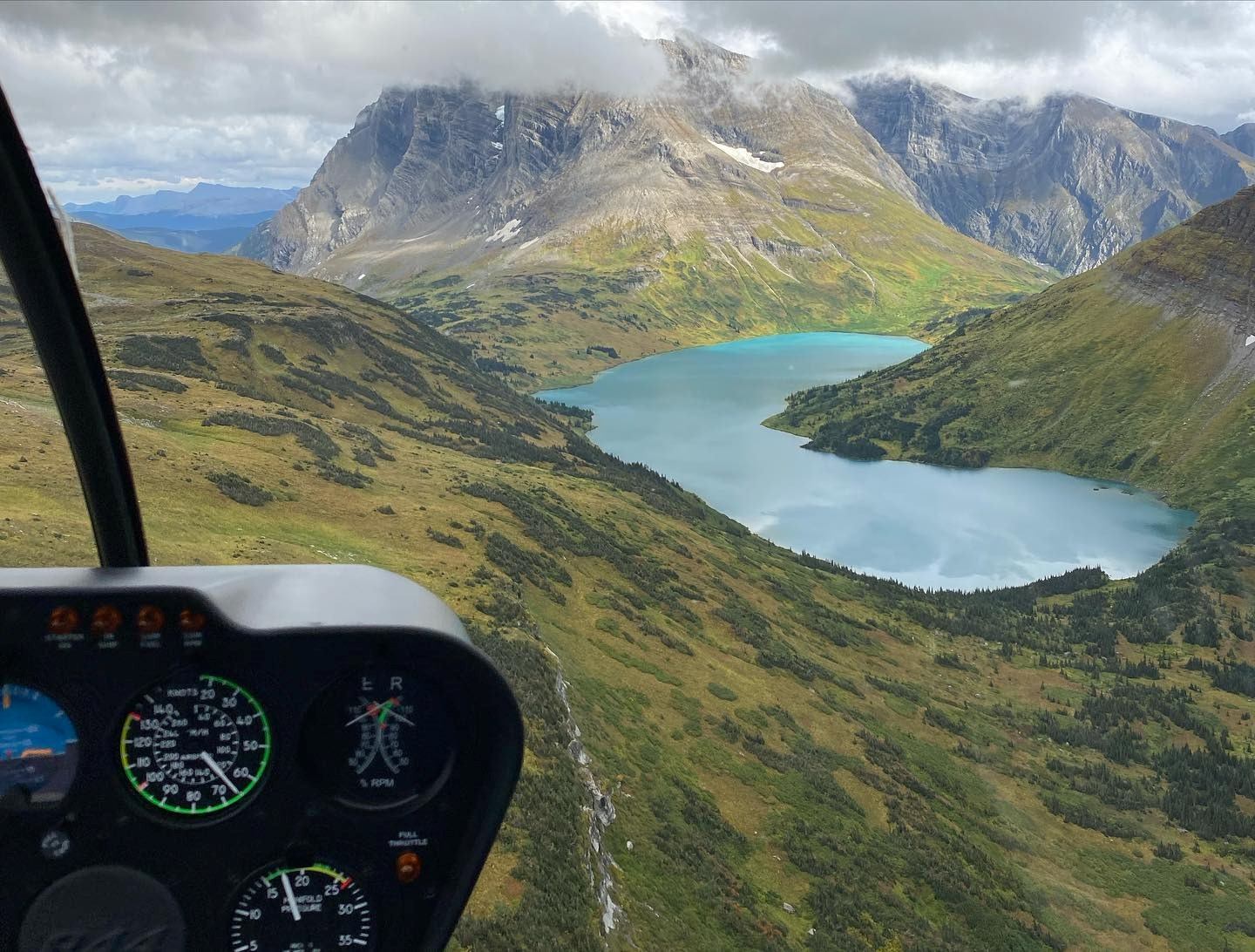 Cockpit view over mountains, valleys, and turquoise lake under partly cloudy sky.
