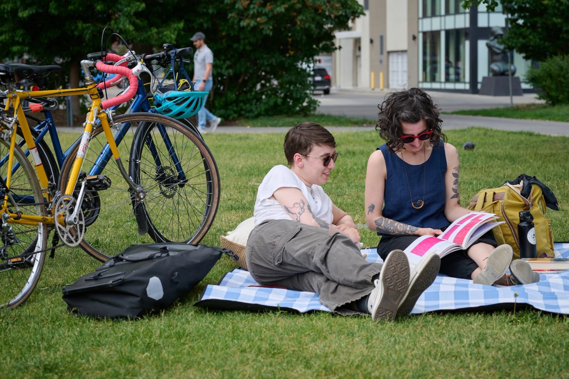 Two people relax on a picnic blanket near bicycles, reading a colourful book together.
