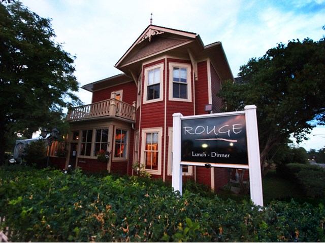 Historic red two-story building with white trim, lush greenery in front. A sign reads "Rouge Lunch • Dinner," evoking a quaint, cozy atmosphere.