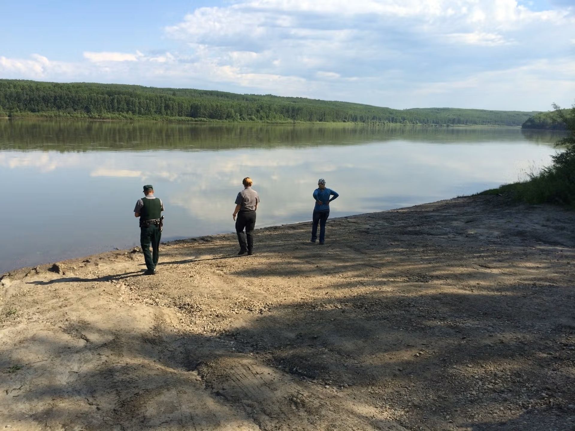 Three visitors stand on a gravel shoreline, looking across calm water bordered by forested hills.