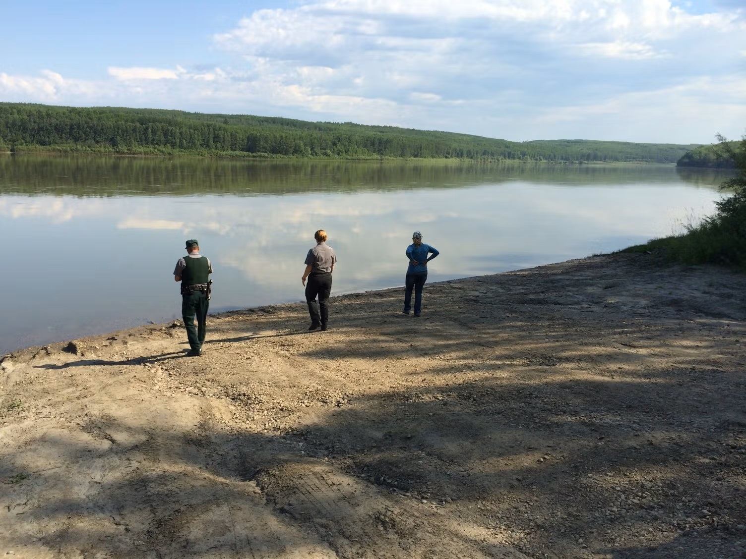 Three visitors stand on a gravel shoreline, looking across calm water bordered by forested hills.