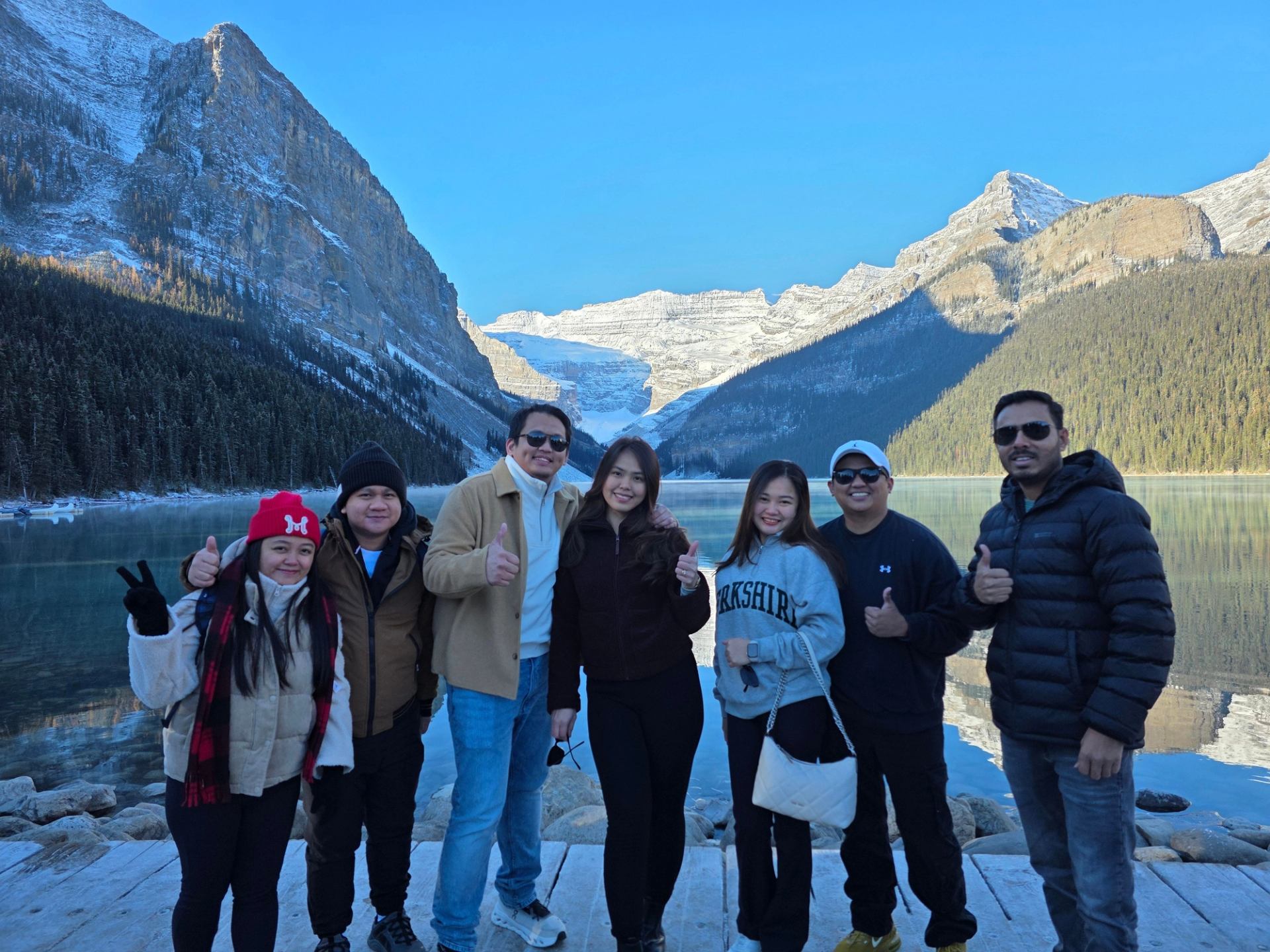 Group of people posing on lakeside boardwalk with snow-capped mountains and forest.