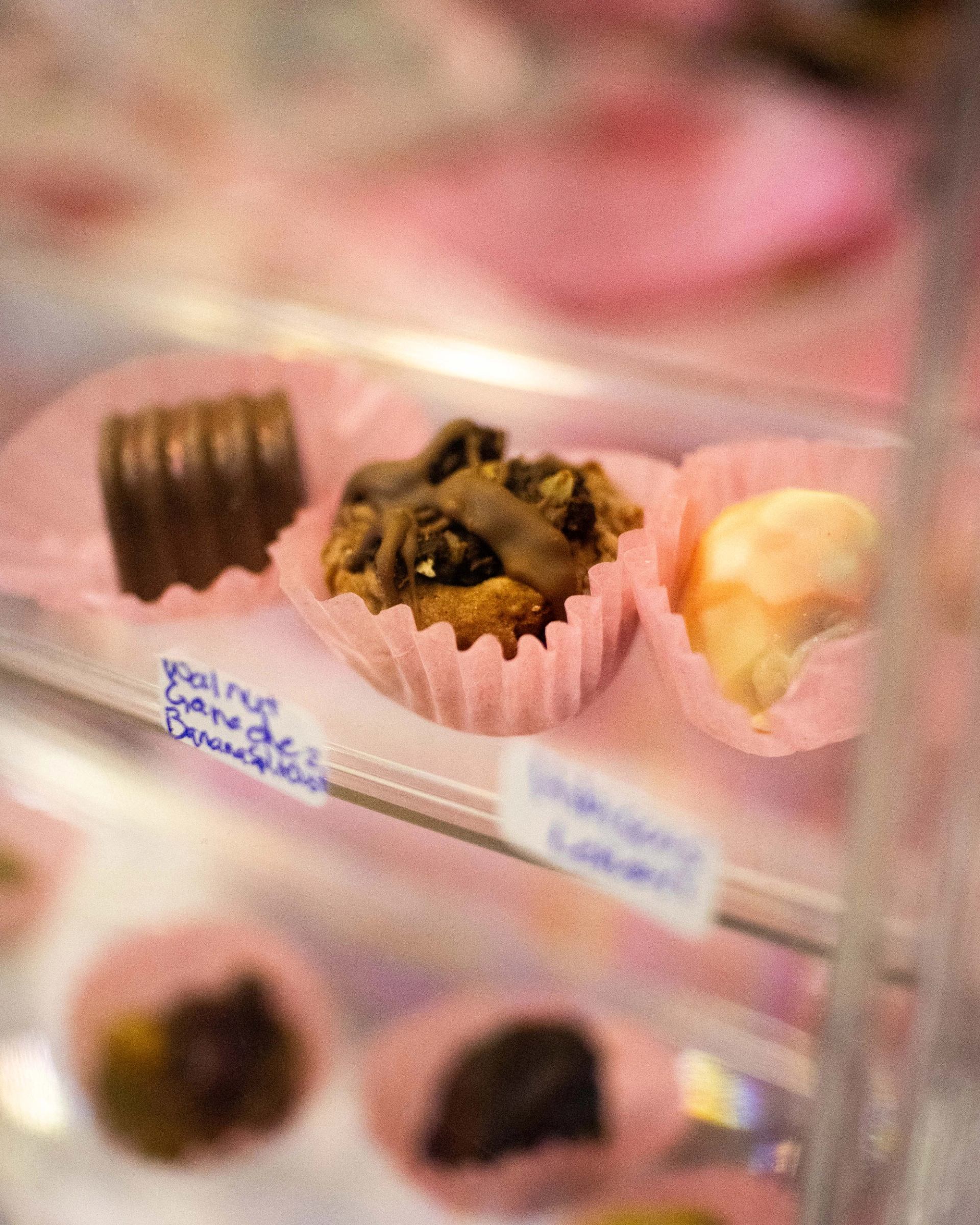 Assorted chocolates in pink paper cups displayed behind glass in a confectionery case.