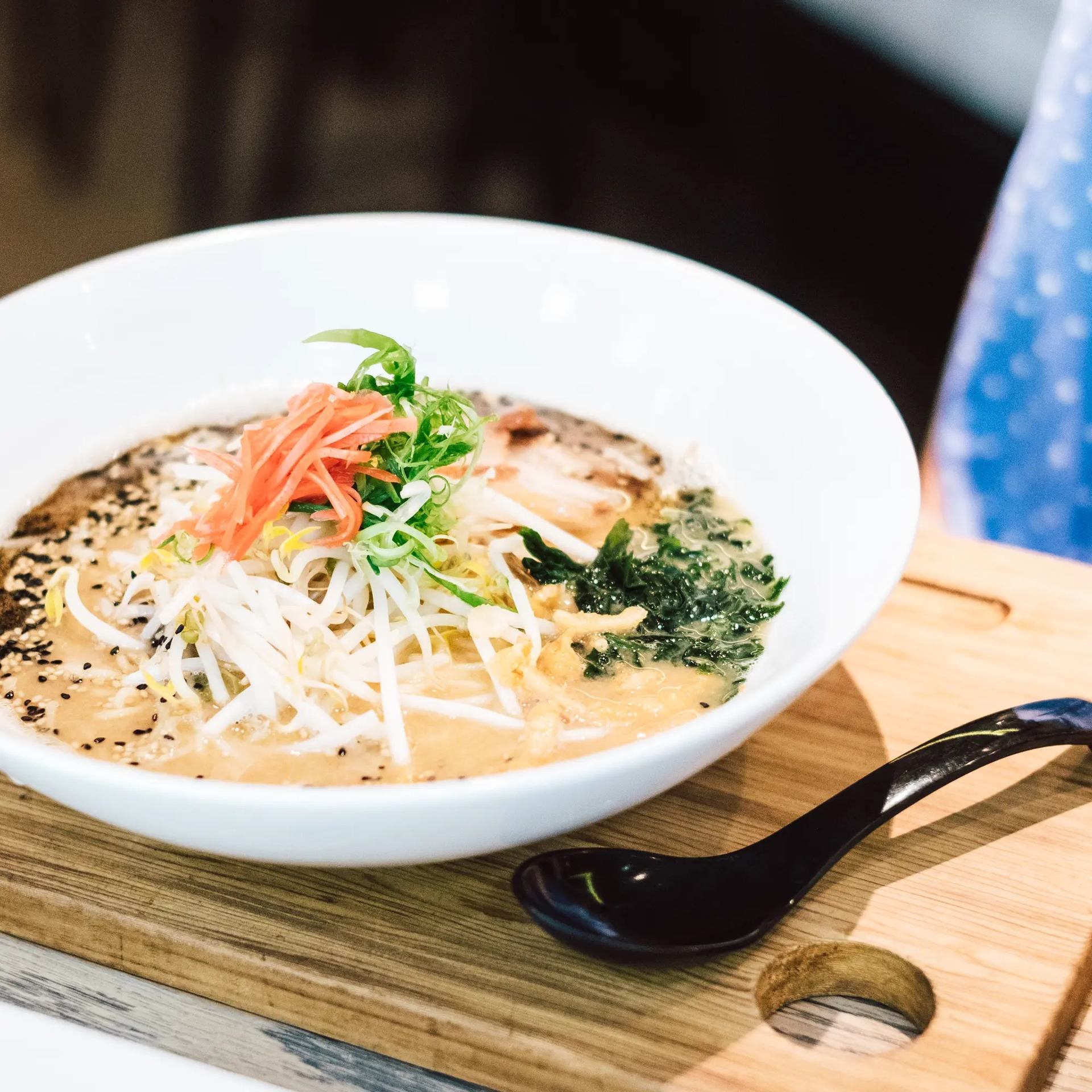Bowl of ramen with toppings on wooden board and black spoon.