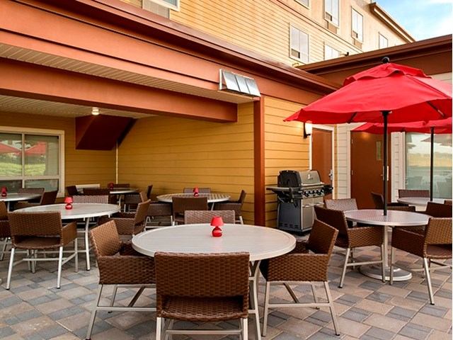 Outdoor patio with tables, chairs, red umbrella, and BBQ grill near yellow building.