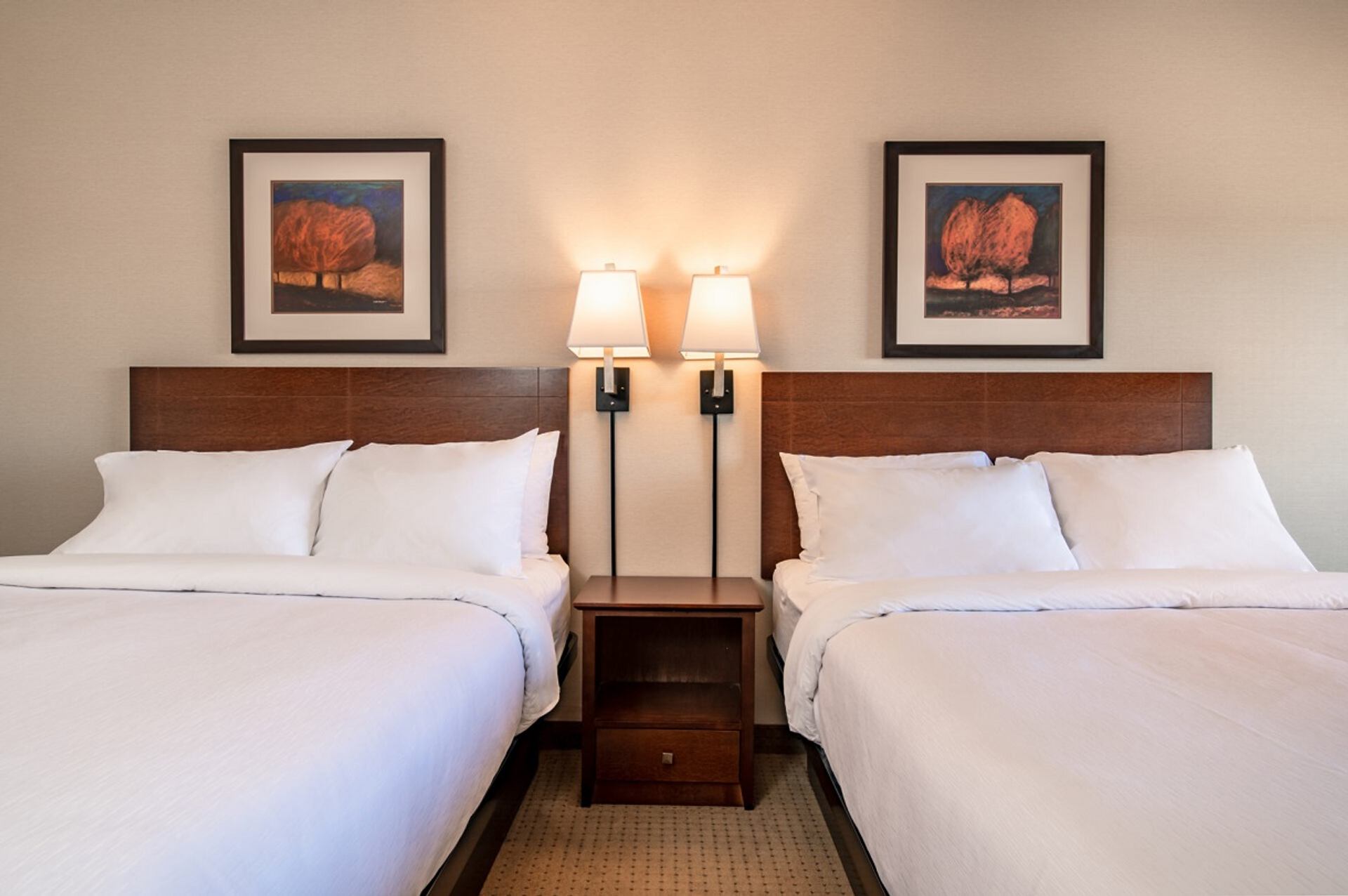 Hotel room with two made beds, shared nightstand, wall lamps and framed art.