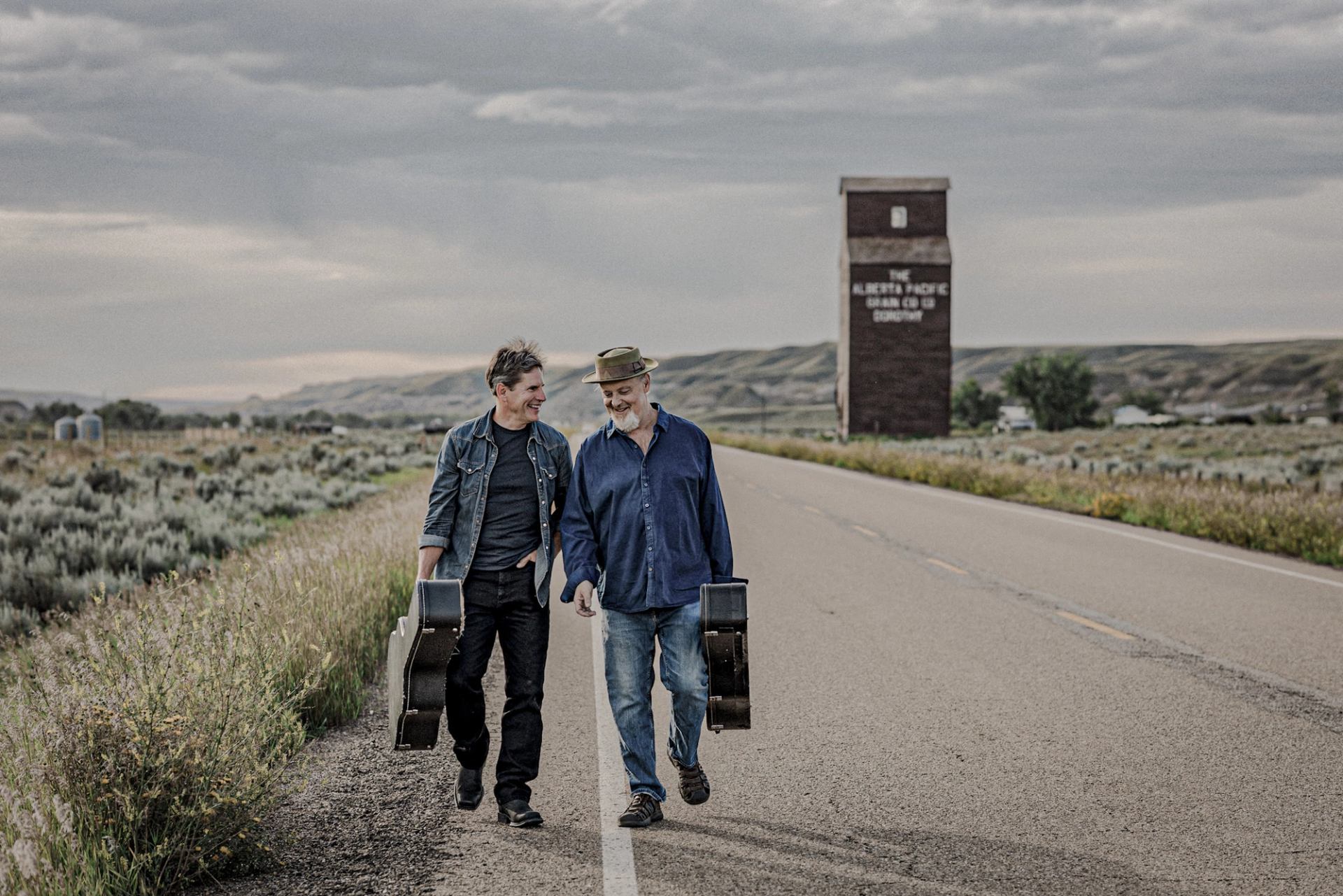 Two people carrying guitar cases walk along rural highway toward grain elevator.