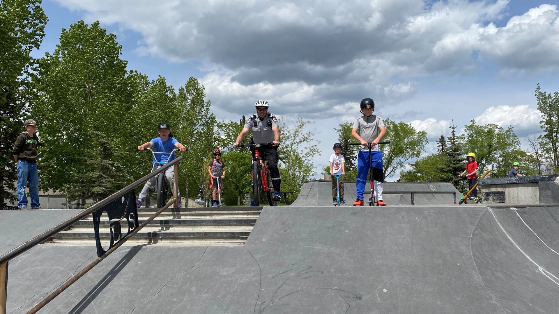 Cyclists and scooter riders wait their turn at the top of a skate ramp.