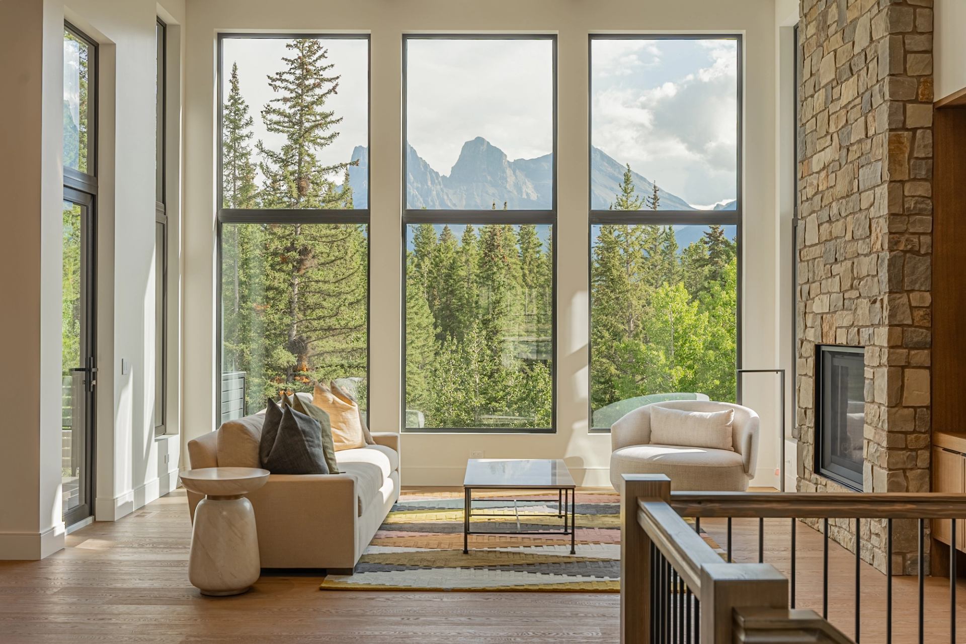 A living room with floor-to-ceiling windows overlooks a forest and mountain landscape, featuring comfortable seating and a stone fireplace.