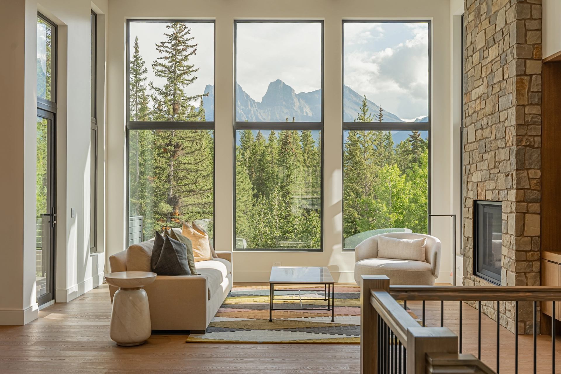 A living room with floor-to-ceiling windows overlooks a forest and mountain landscape, featuring comfortable seating and a stone fireplace.
