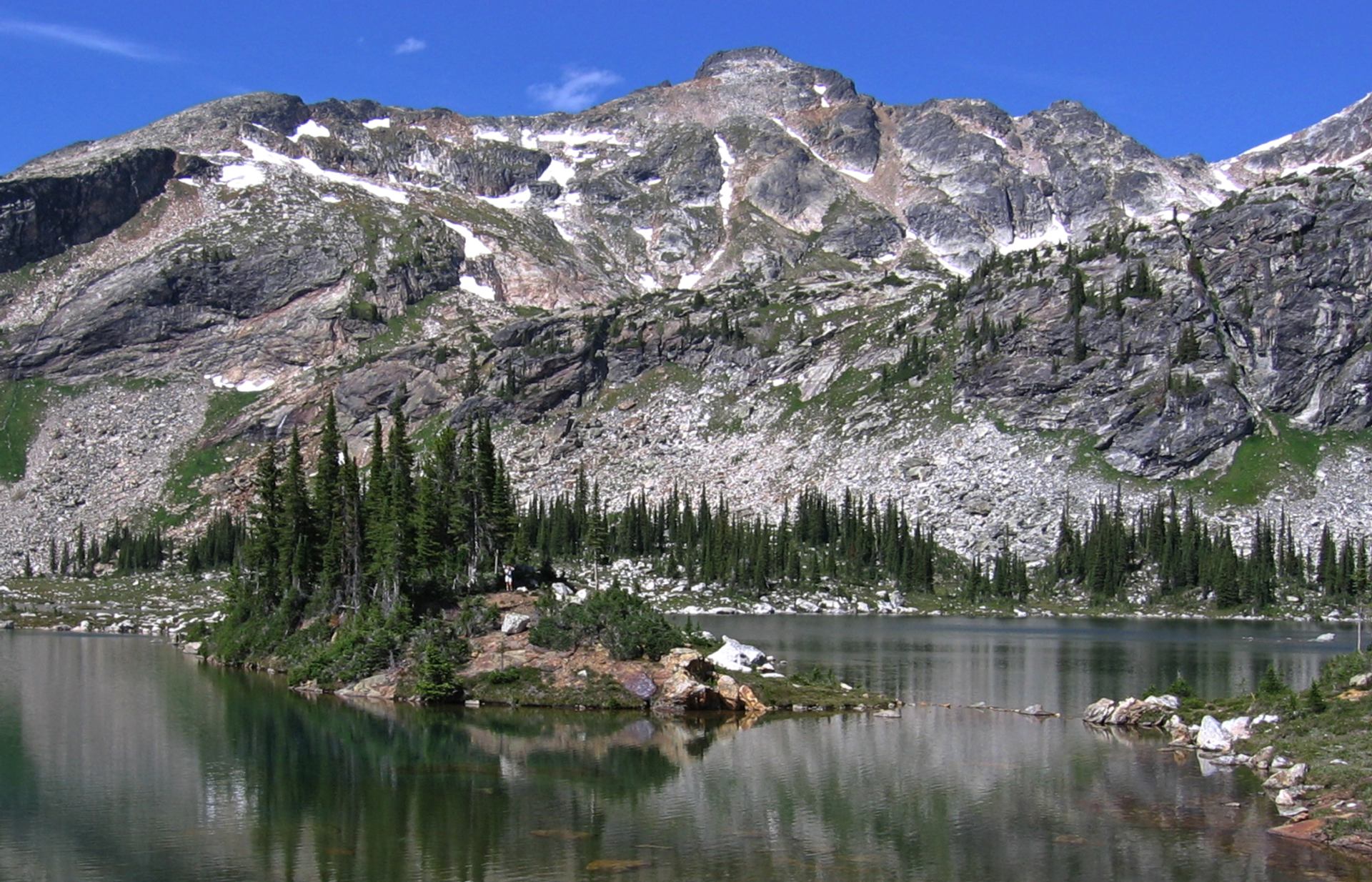Alpine lake reflects rocky mountain slope with scattered snow and dense evergreen trees.