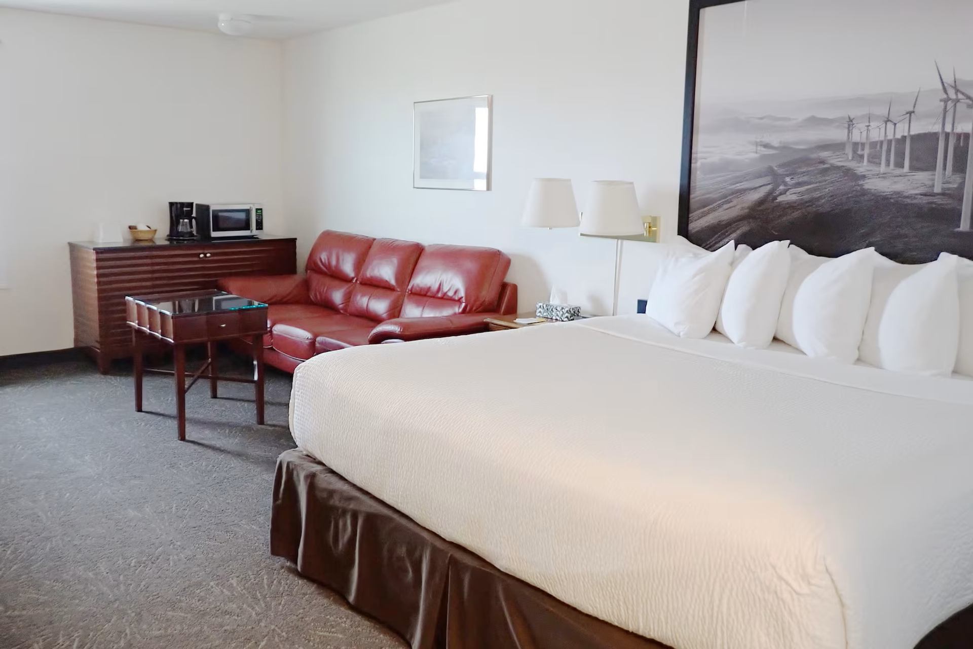 Hotel room with a large bed, red sofa, coffee station, wall lamps, and a mural featuring wind turbines.
