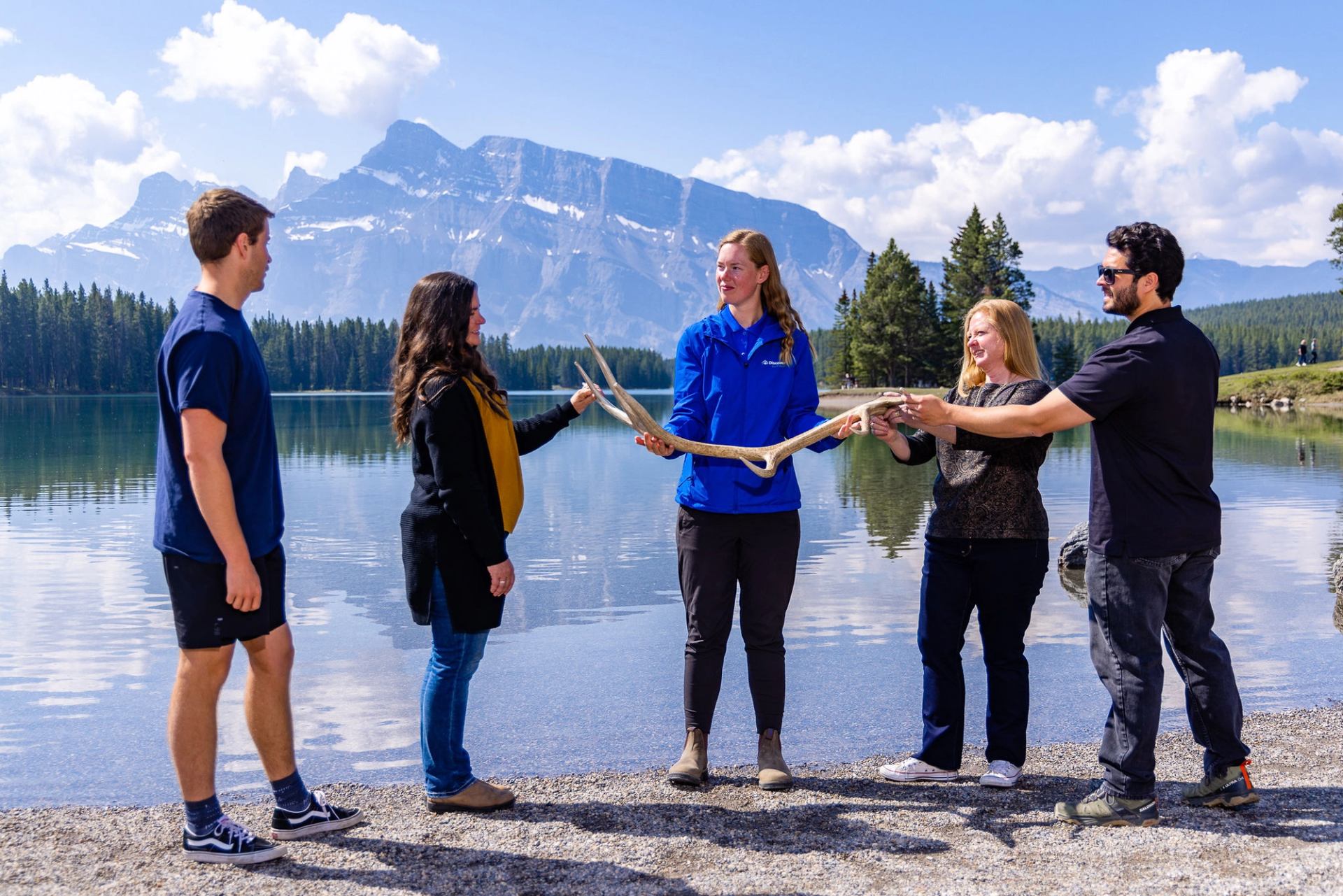 Group of people holding large antlers beside a calm mountain lake.