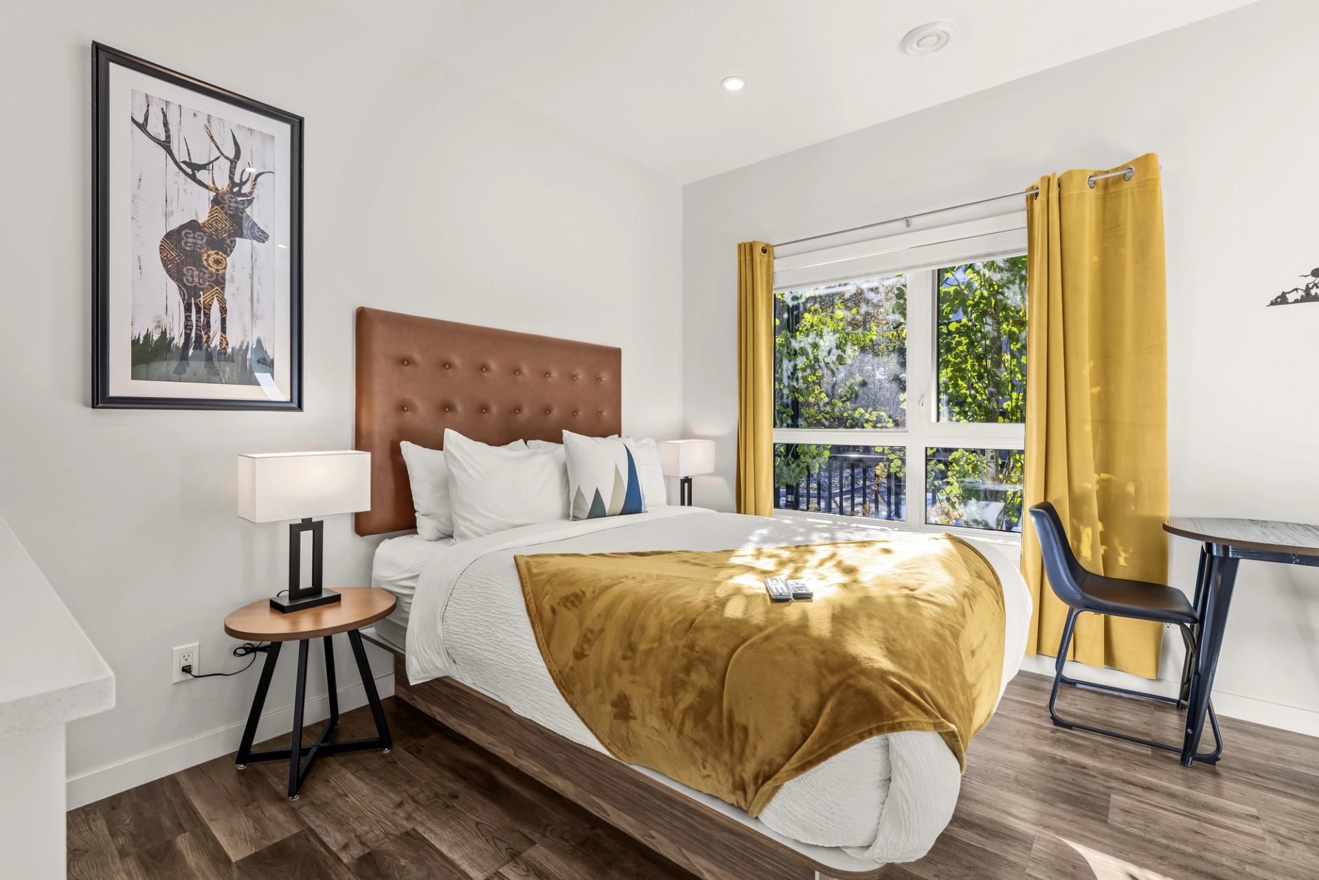 Bedroom with mustard curtains, matching blanket, framed deer artwork and window overlooking trees.