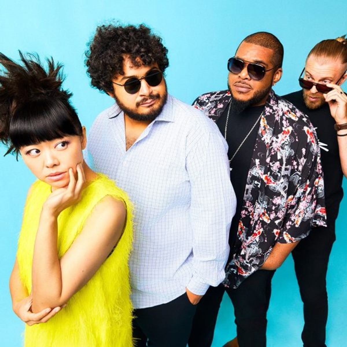 Four people in bright clothing and sunglasses pose playfully against a blue studio backdrop.