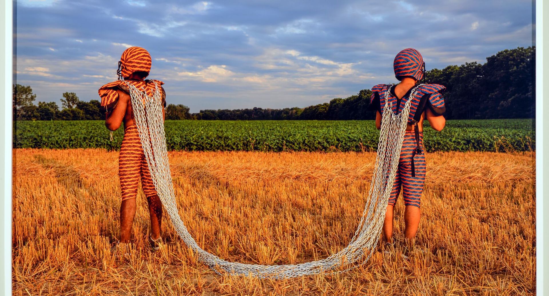 Two costumed figures stand in a harvested field, linked by long draped metal chains.