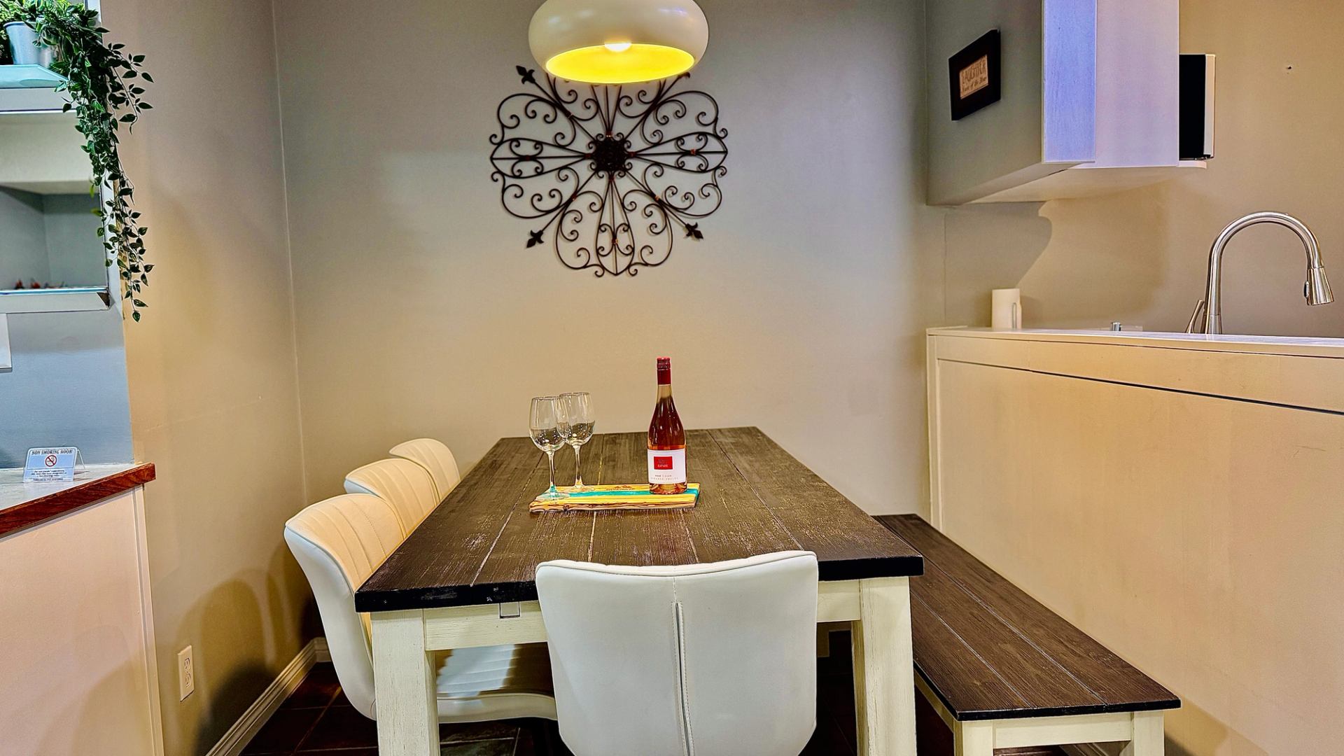 Dining table with white chairs, bench, wine bottle and glasses beside a kitchen.