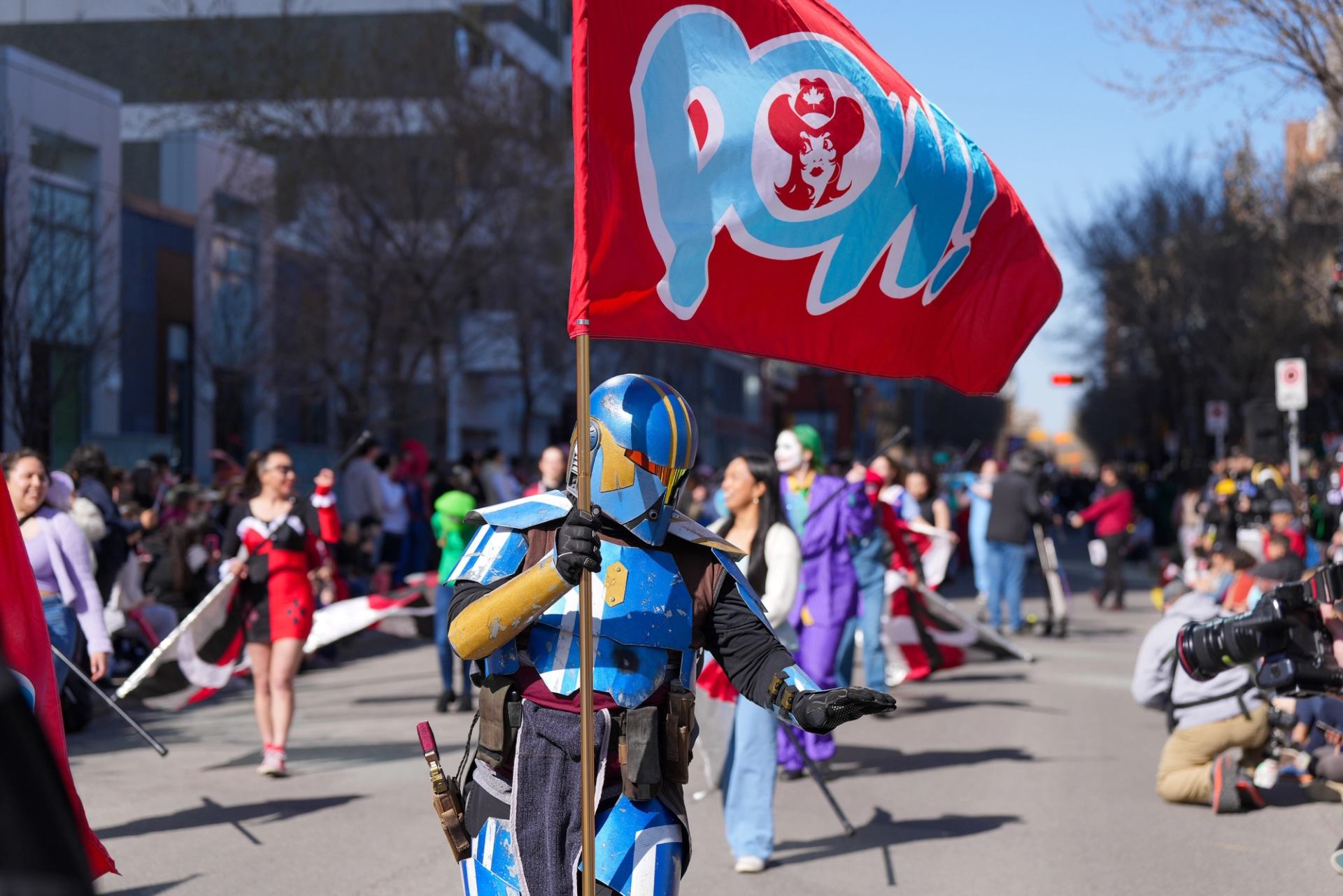 Armoured cosplayer carries large red POW flag while walking in crowded city street parade.