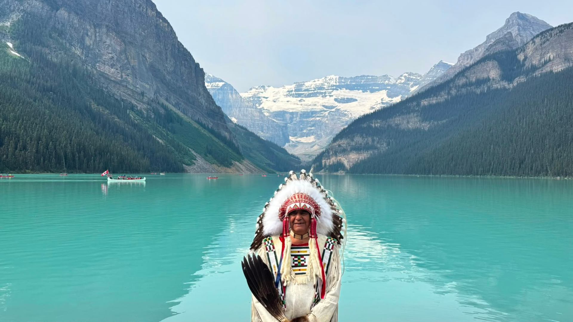 Person in a feathered headdress stands before a turquoise lake and snow-capped mountains.