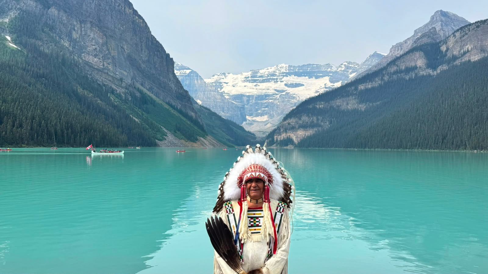 Person in a feathered headdress stands before a turquoise lake and snow-capped mountains.
