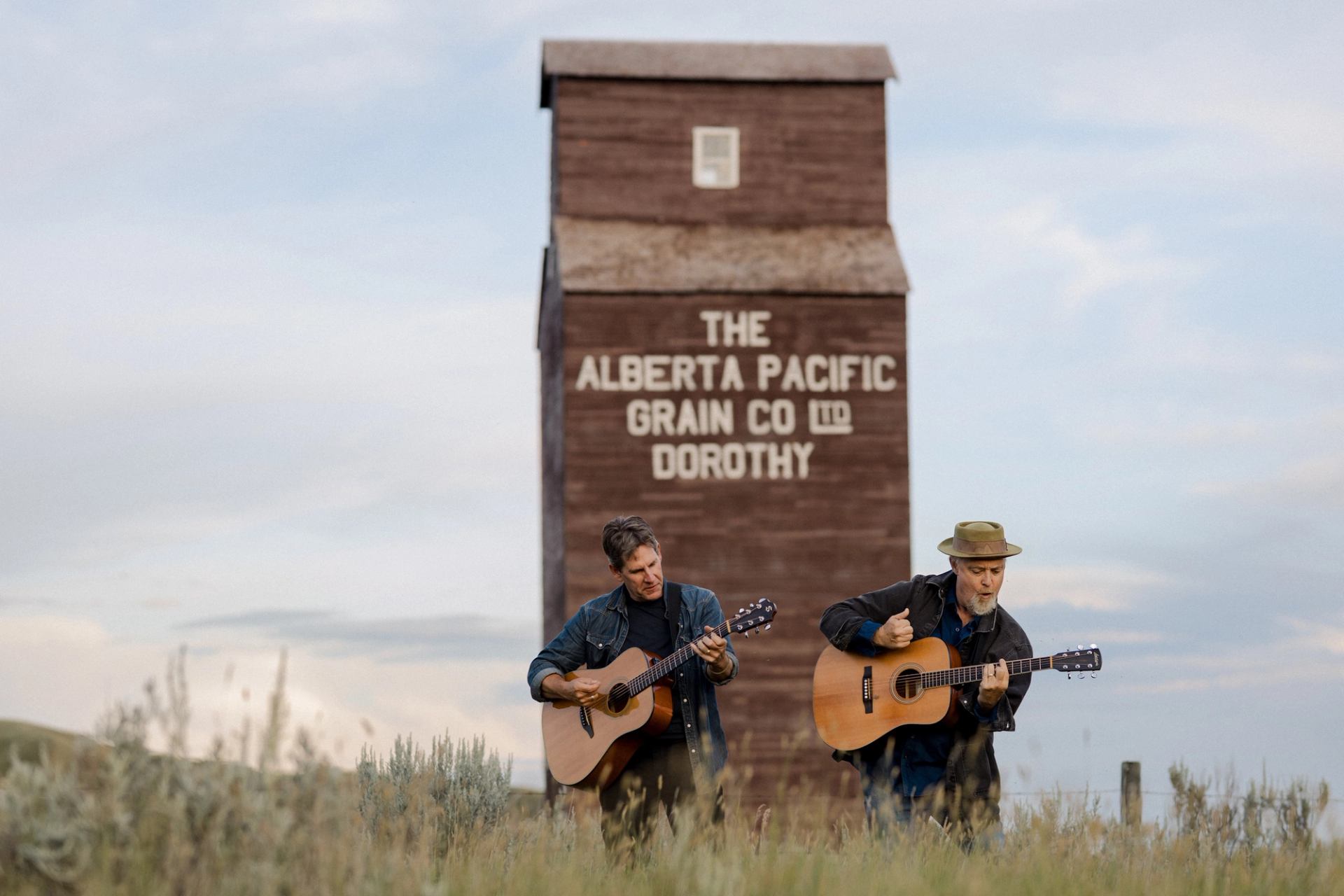 Two people play acoustic guitars in tall grass before Alberta Pacific Grain Co elevator.