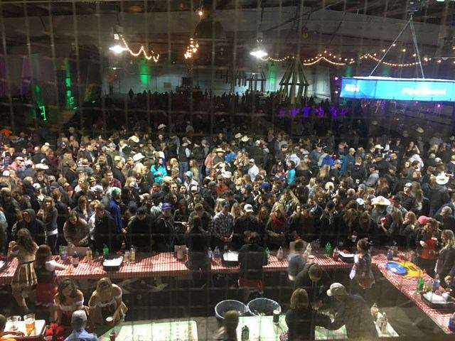 Large indoor crowd in cowboy hats gathers near food tables under string lights.