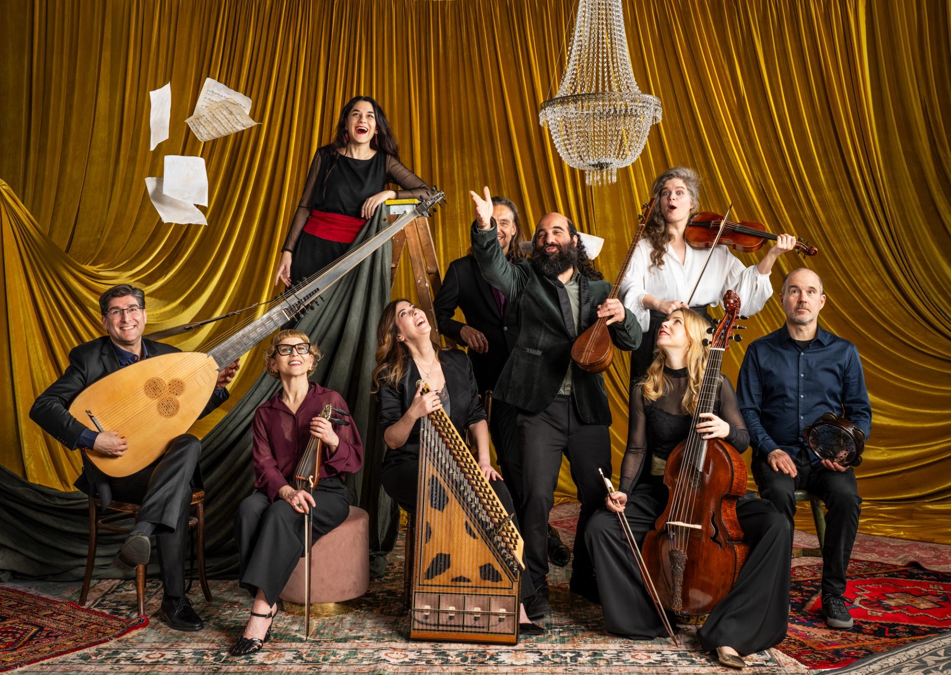 Ensemble of musicians with historical instruments pose before gold curtain and chandelier.