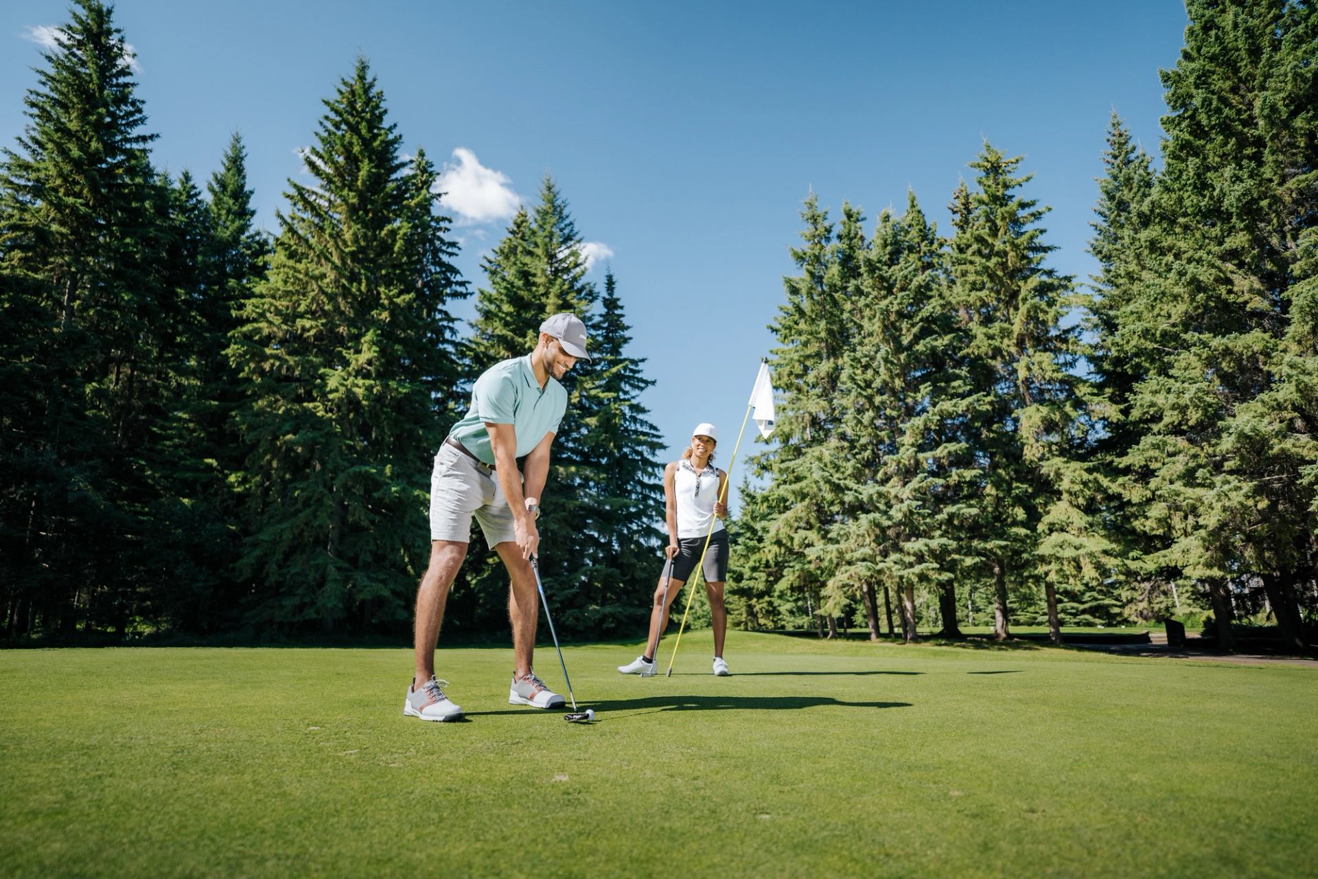 wo golfers on a lush green putting area surrounded by tall pine trees