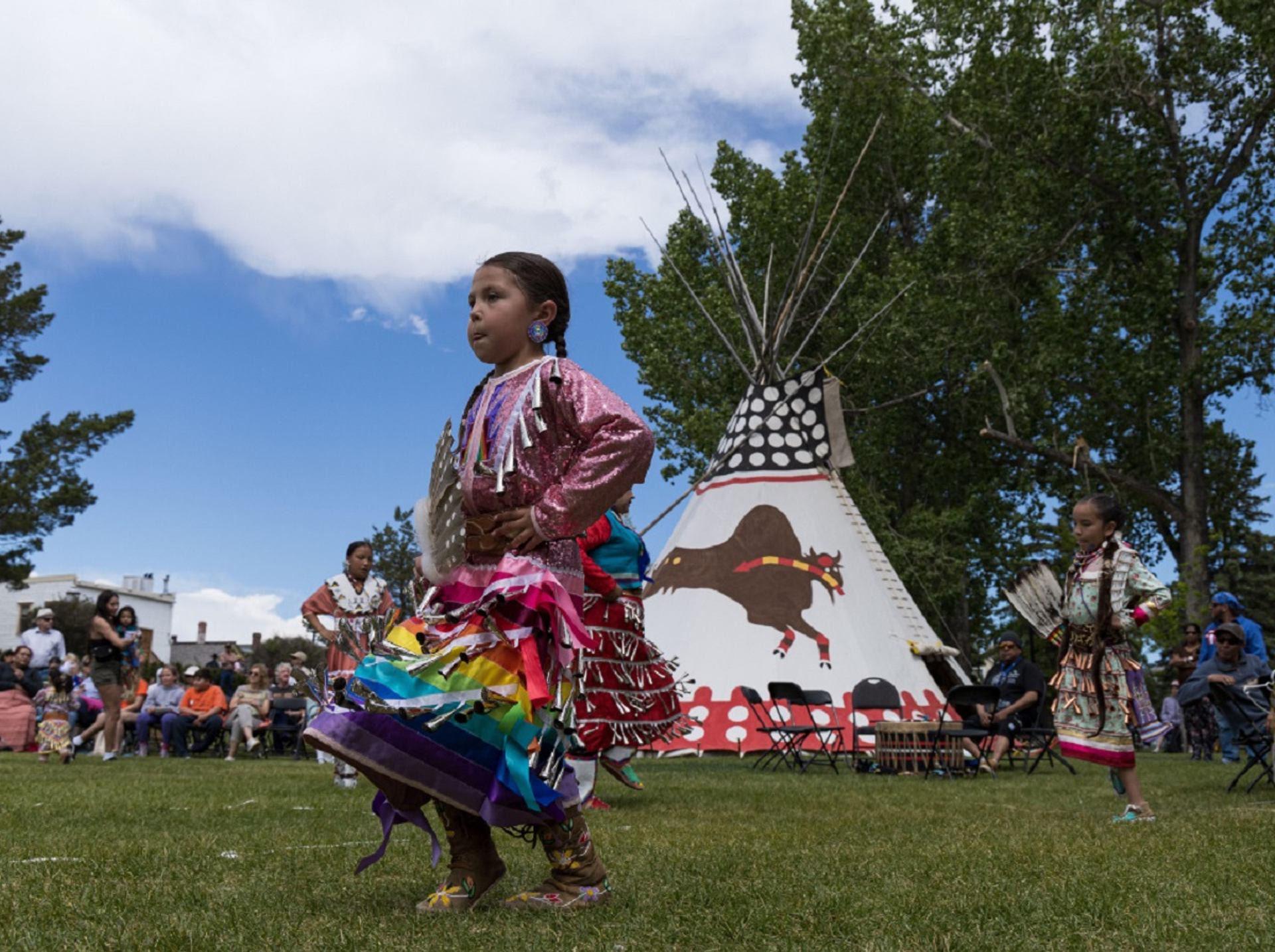 Children in colourful regalia dance on grass near painted tipi, with spectators watching.