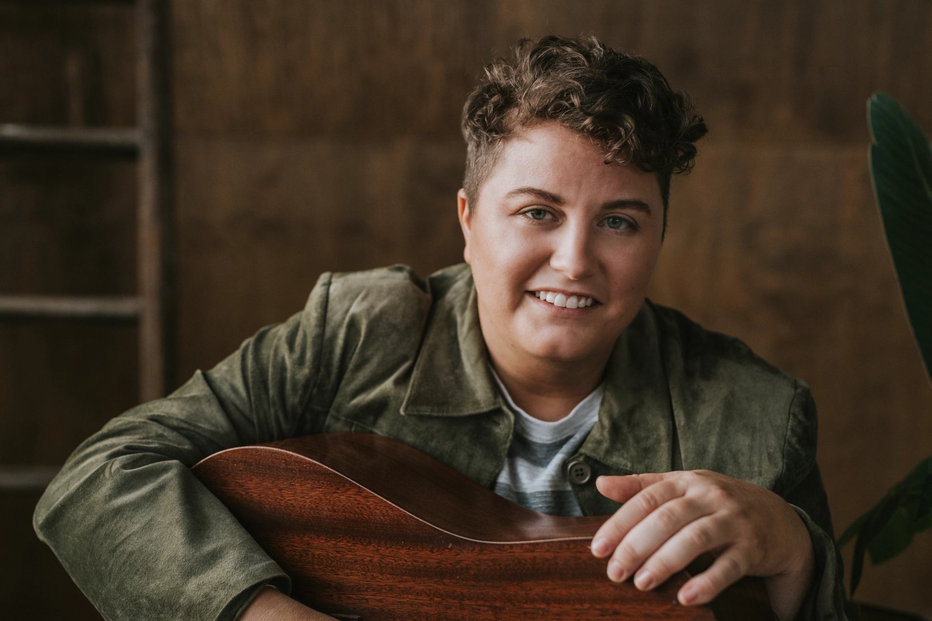 Person in green jacket holding acoustic guitar, seated indoors against wooden wall.