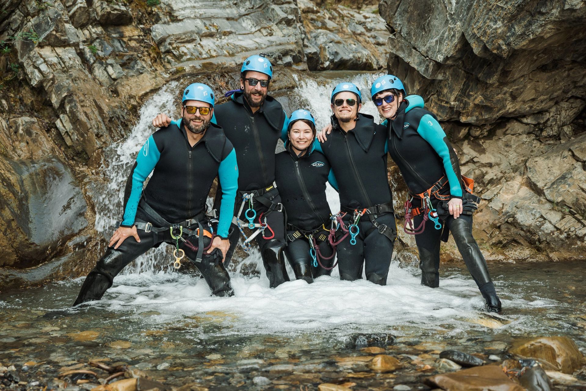 Six people in wetsuits, helmets, and harnesses stand in a stream with a waterfall, ready for canyoning.