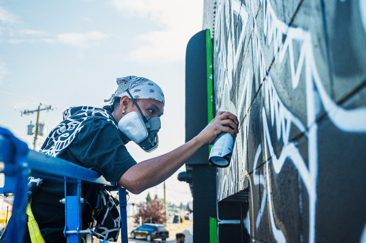 Person in bandana and mask spray-paints graffiti art on a wall from an elevated platform.
