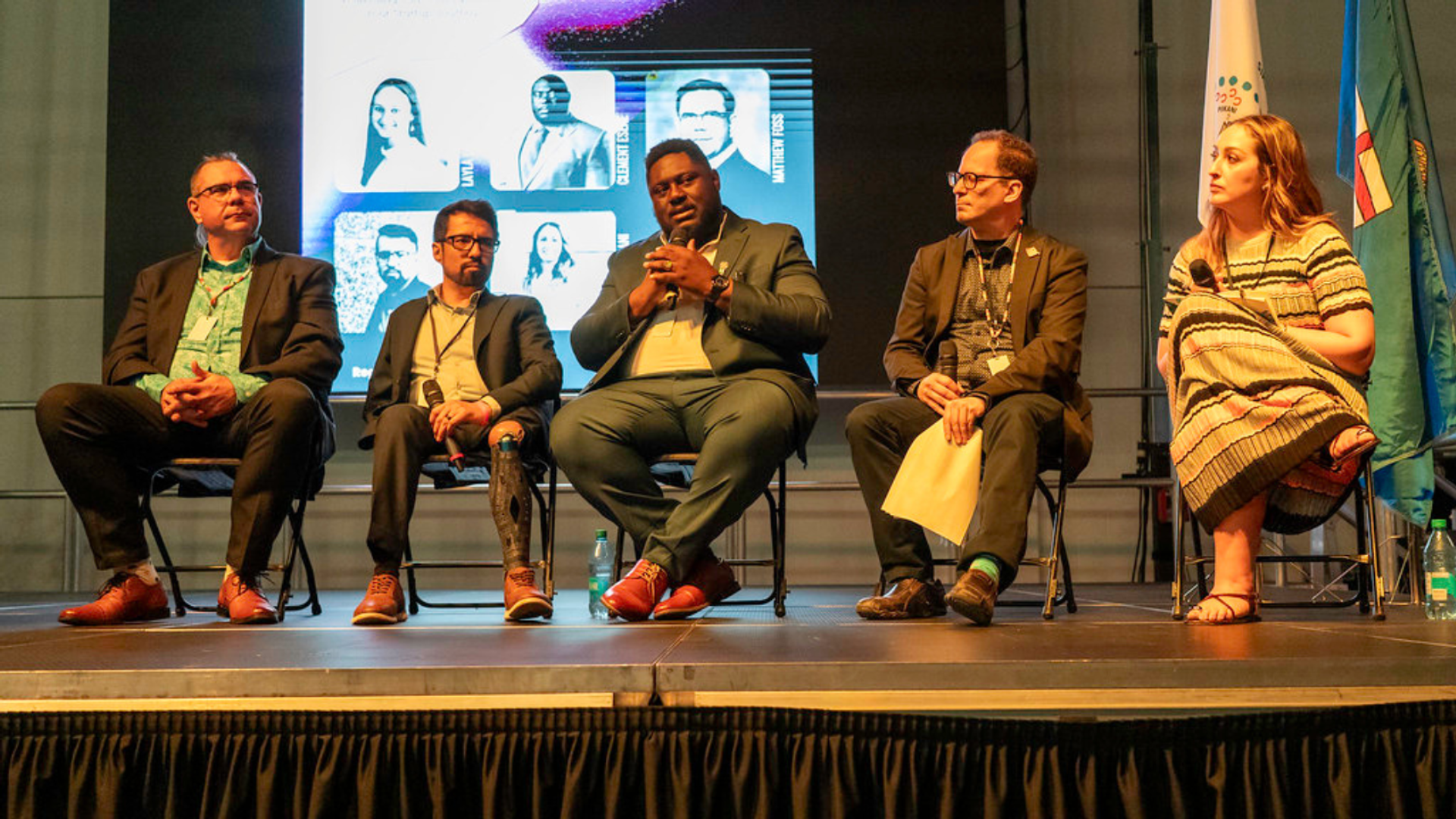 Panel of speakers seated on stage in front of a large screen during a conference session. 