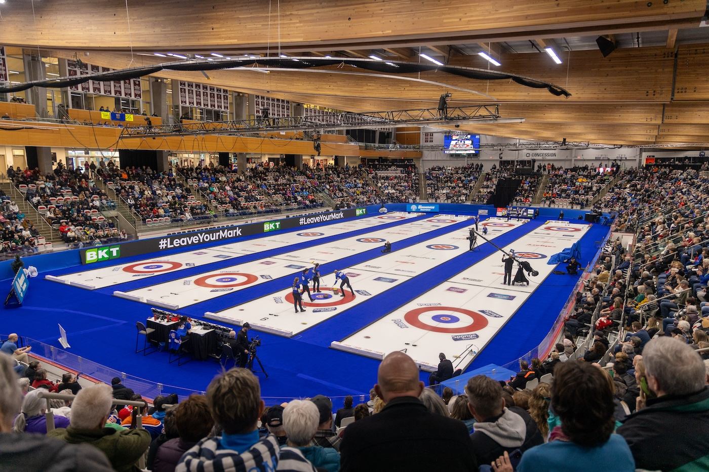 Panoramic view of a curling arena filled with spectators watching multiple curling matches in progress.