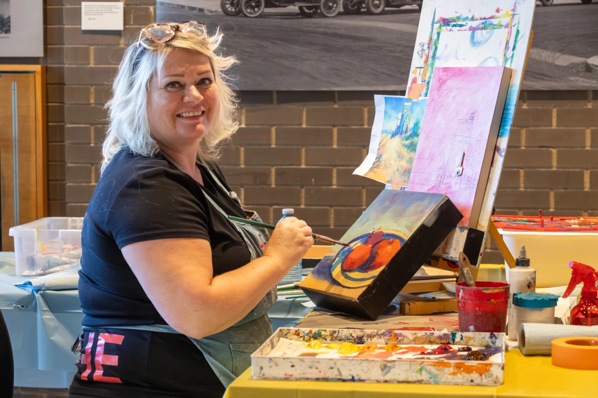 Artist in apron paints colourful fruit still life on canvas at cluttered studio table.