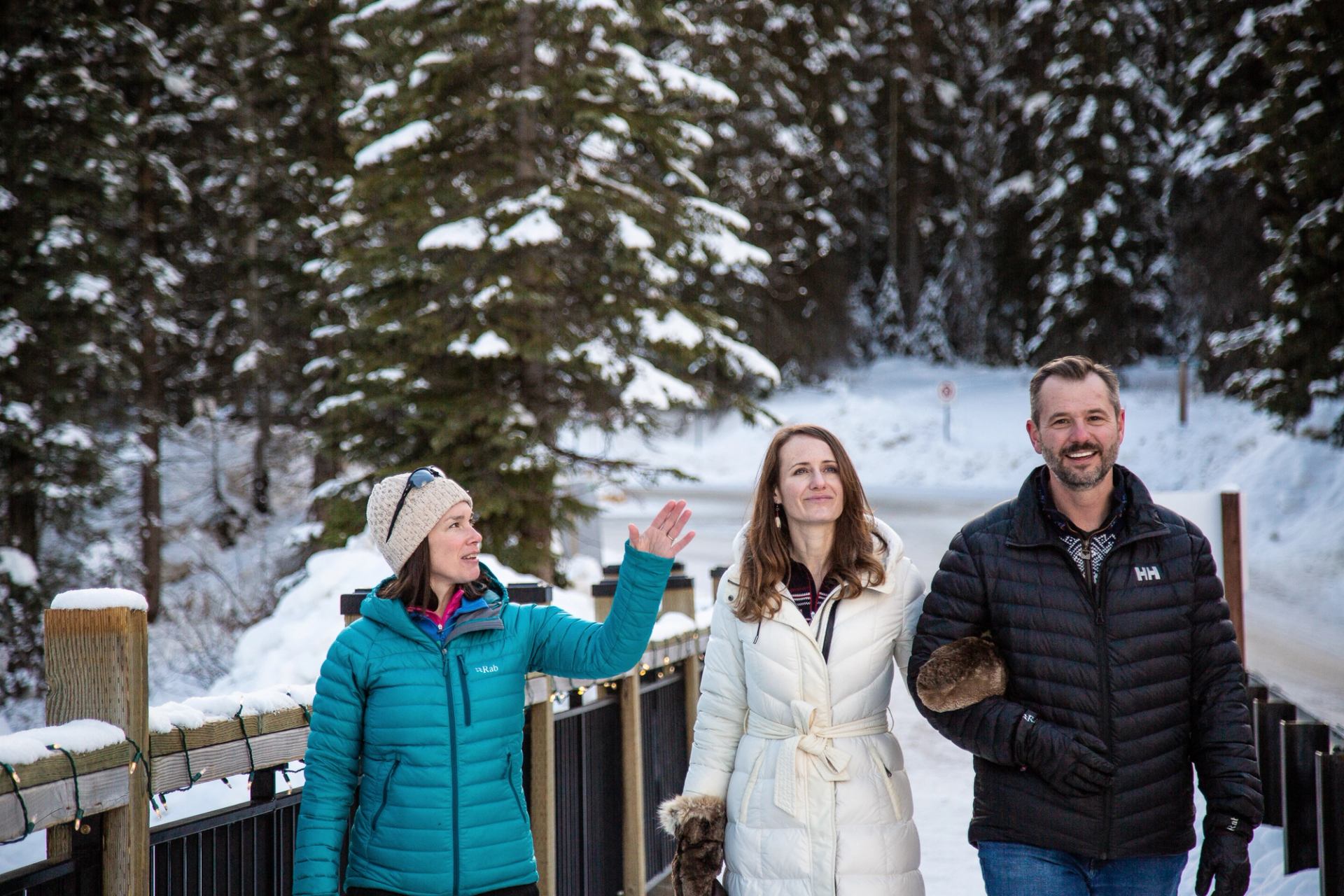 Three people walking on a snowy path surrounded by pine trees.