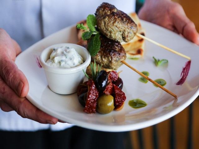 A chef holds a plate with two grilled meat skewers garnished with mint, a white dip, olives, sun-dried tomatoes, and flatbread.