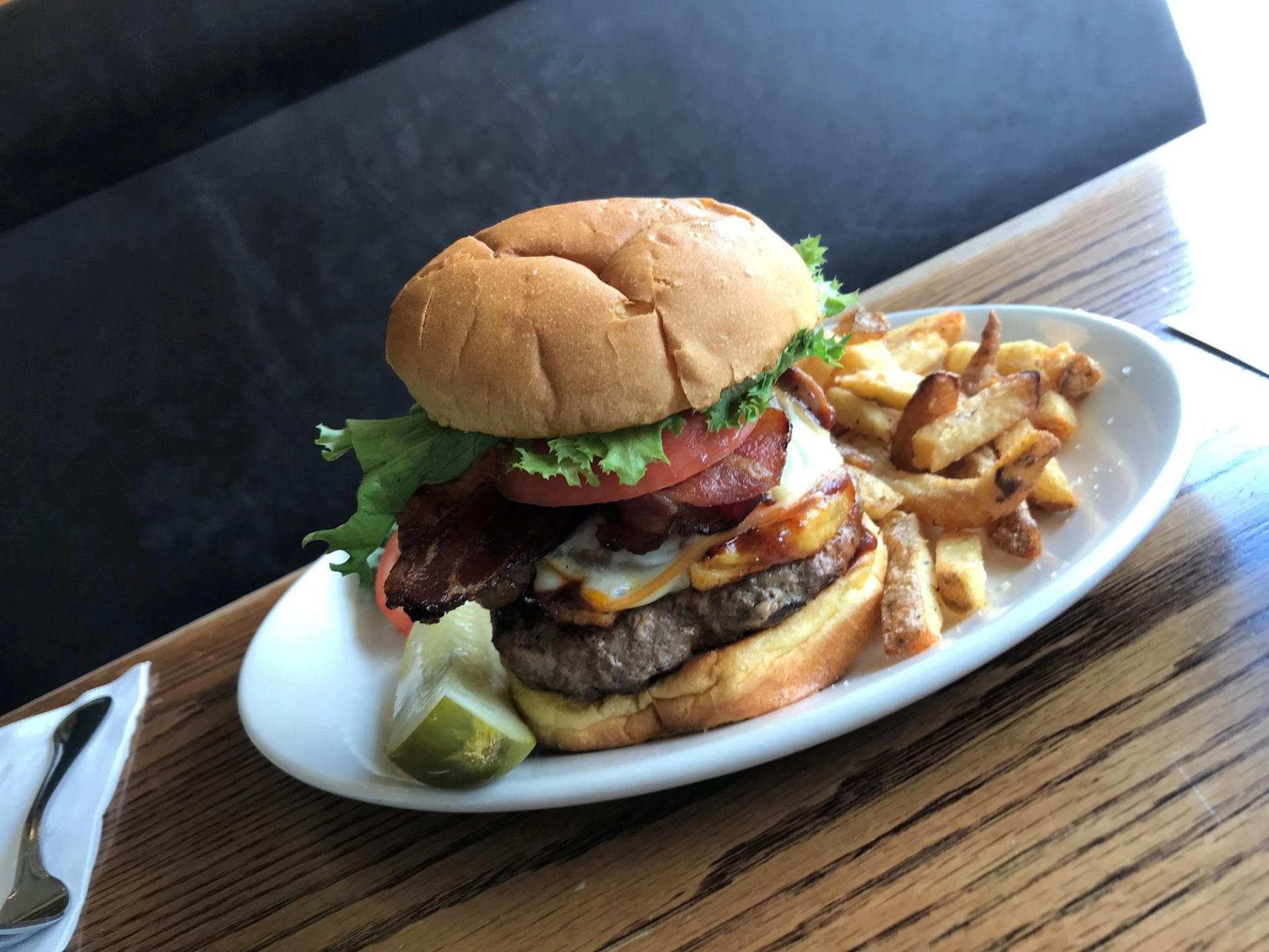 Bacon cheeseburger with fries and a pickle on a white plate on a wooden table.