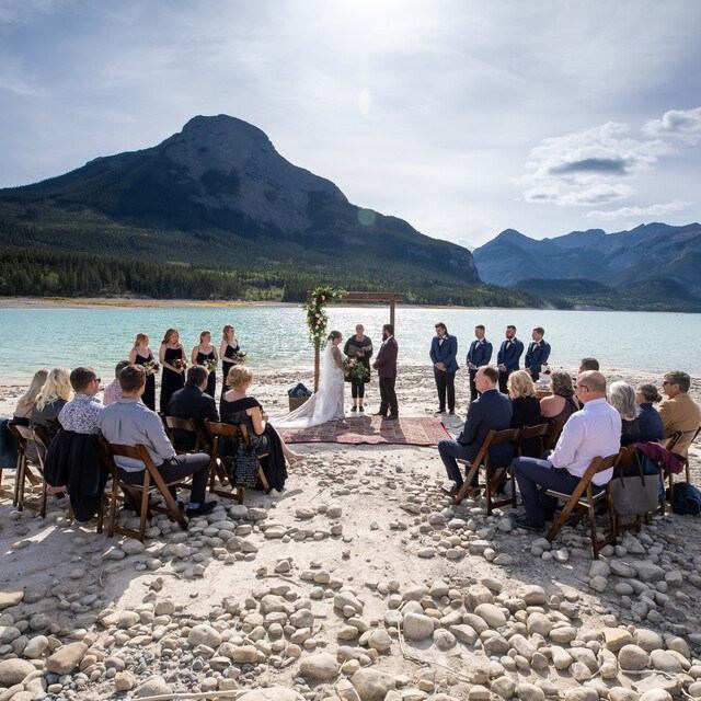 Outdoor wedding ceremony beside a turquoise lake with mountains, floral arch, and seated guests.