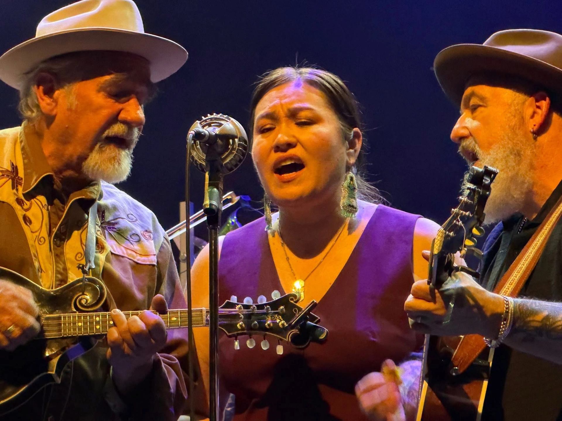 Three musicians sing into a vintage microphone while playing string instruments onstage.