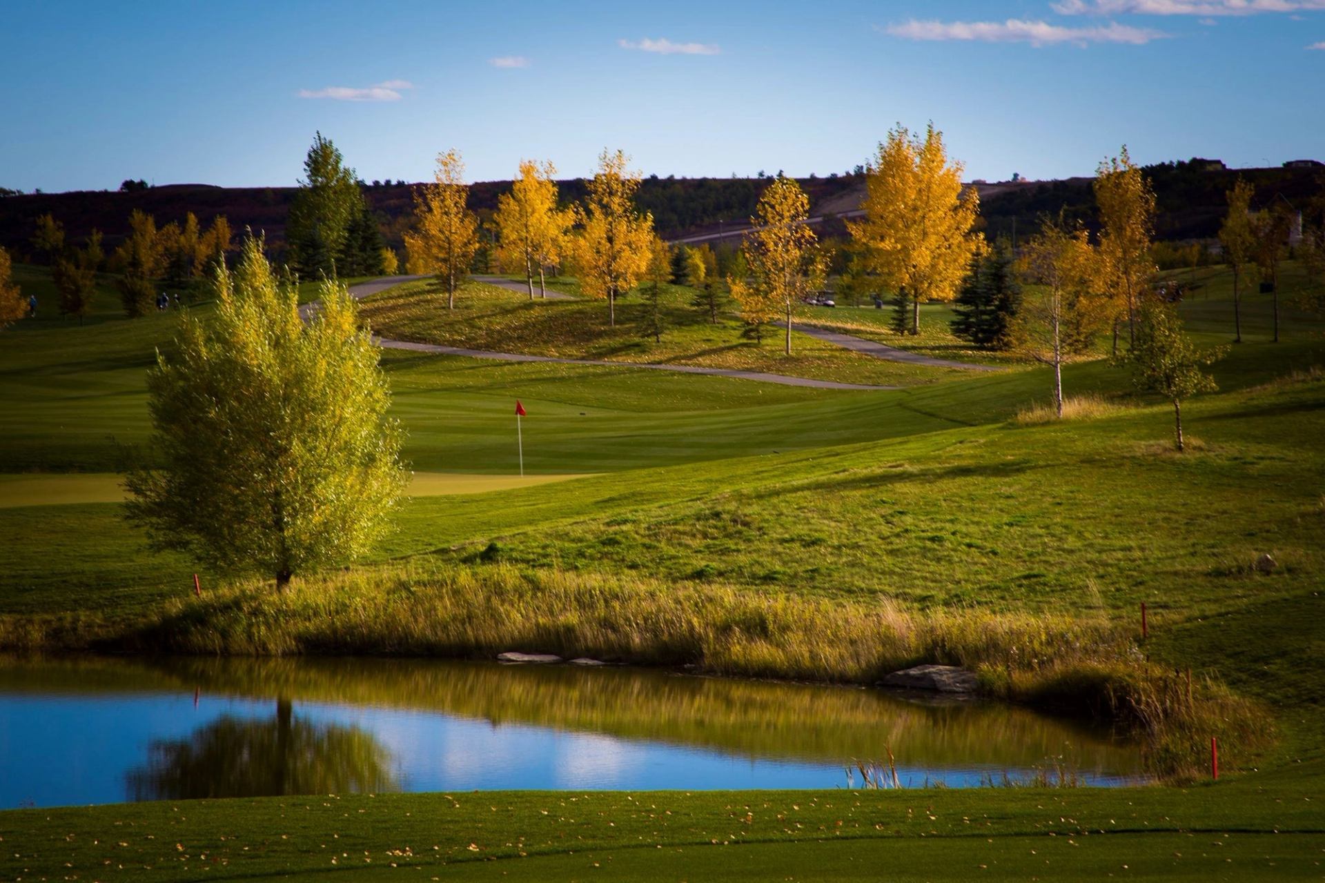 Golf course with a scenic pond, trees with autumn colors, and a red flag marking the hole.