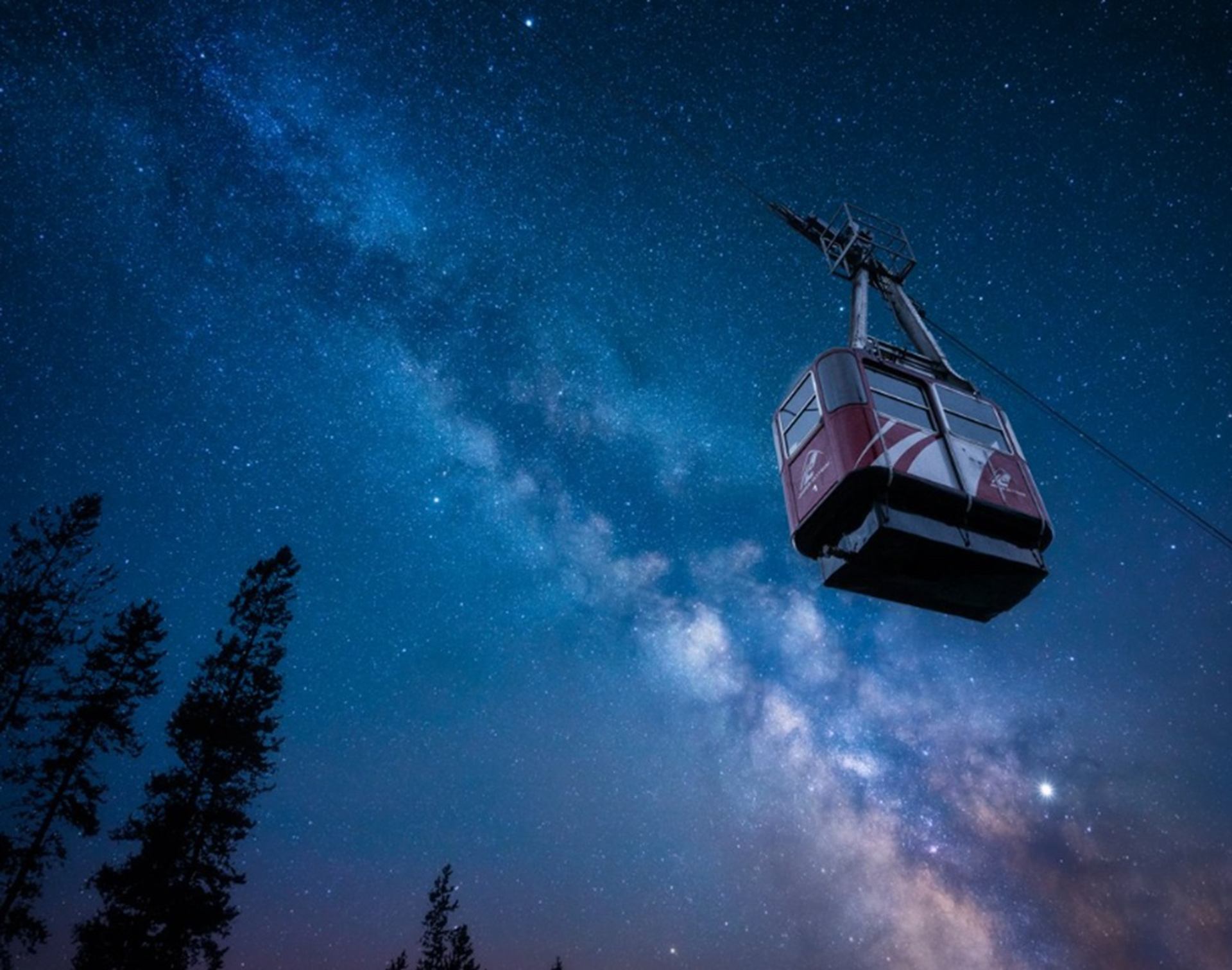 Red gondola hangs above silhouetted trees under bright Milky Way night sky.