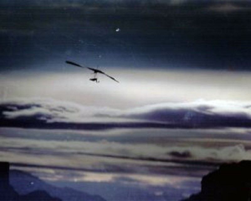 Hang glider above cloud layer with distant mountain ridges below.