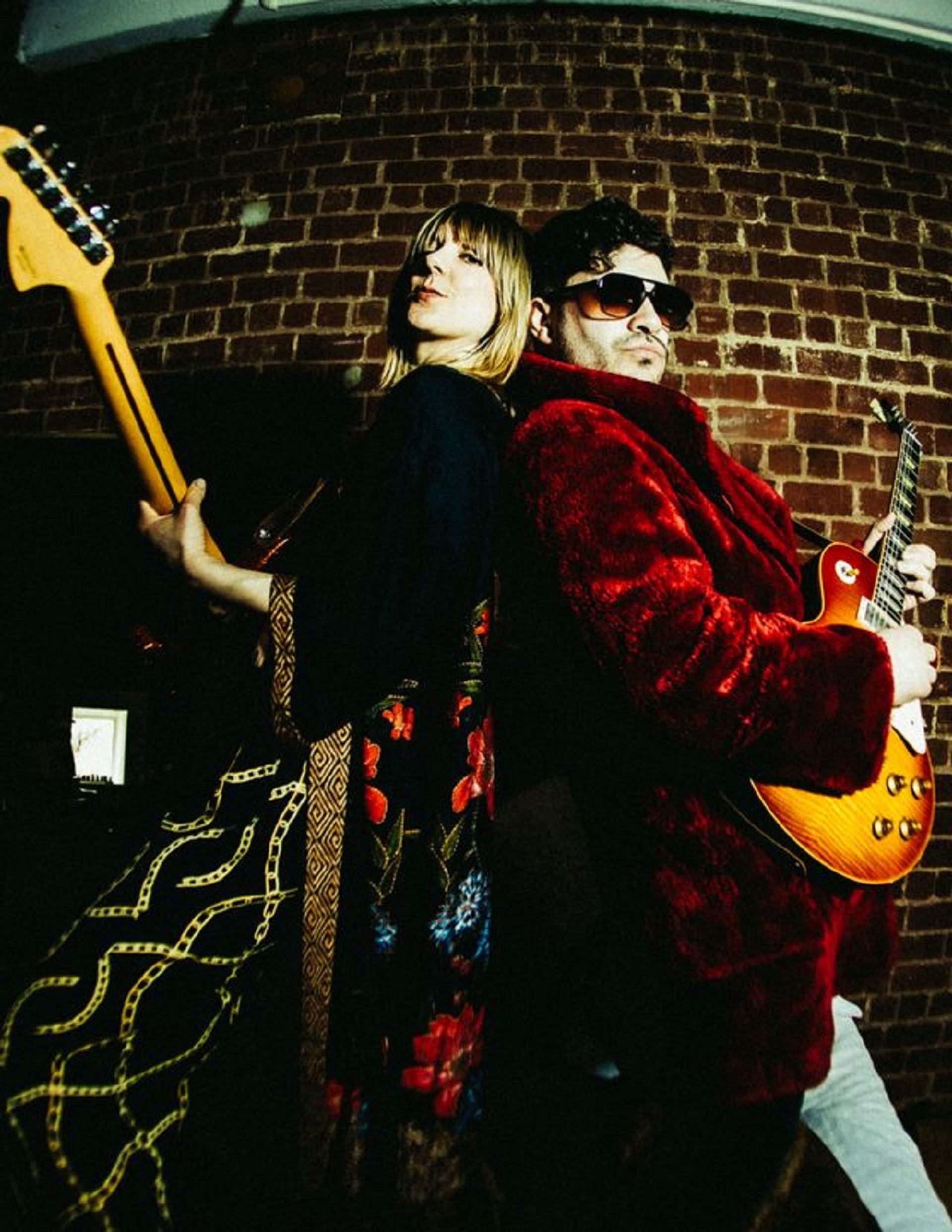 Two guitarists stand back to back against a brick wall, holding electric guitars.