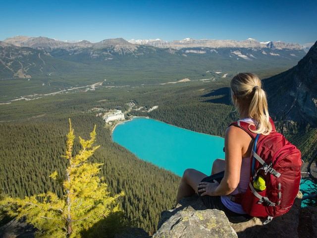 Person with red backpack sits on rocky ledge overlooking turquoise lake and mountains.