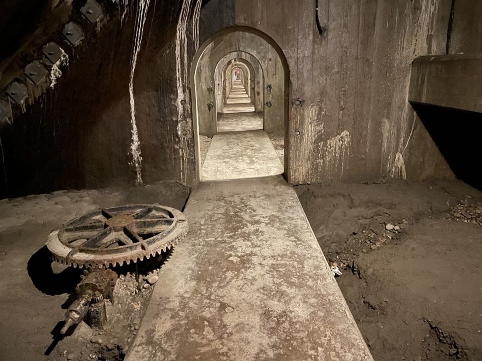 Concrete underground tunnel with repeating archways, exposed pipes and a large rusty gear mechanism.